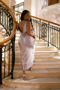 Elite curvy black escort in as long silk dress climbing up a luxurious staircase